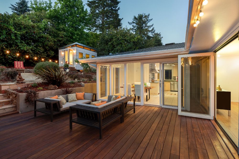 Modern home exterior with illuminated deck, seating area, and open glass doors at dusk
