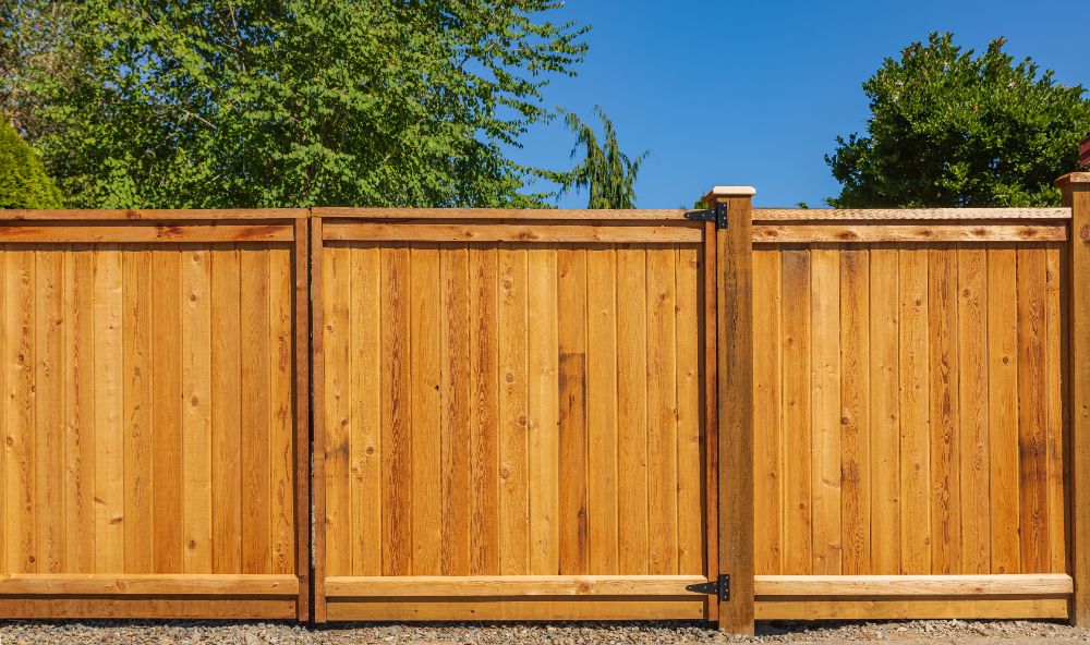 Wooden privacy fence with three panels and metal hinges against green trees and blue sky